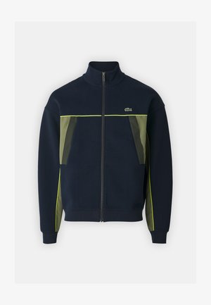 Marineblauwe ritsjack met hoge kraag, groene accenten op zakken en zijkanten, en reflecterende gele piping. Heeft een klein logo op de borst.