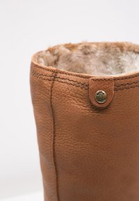 Close-up van een bruin leren laars met zichtbare stiksels, nepbontvoering en een ronde metalen logo-knop dicht bij de bovenrand.