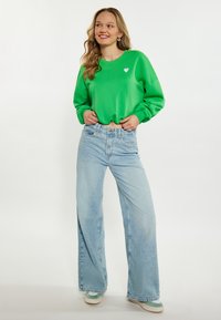 Zielony bawełniany sweatshirt z małym białym sercem, w połączeniu z jasnoniebieskimi dżinsami o wysokim stanie i luźnym kroju oraz trampkami z zielonymi akcentami.