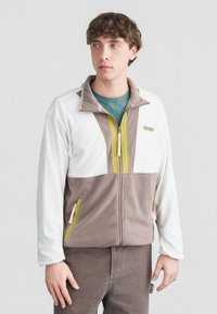 Νέος άνδρας που φοράει φλις ζακέτα με φερμουάρ σε λευκό και μπεζ με πράσινες λεπτομέρειες, στέκεται μπροστά σε ένα απλό φωτεινό φόντο, κοιτώντας προς τα αριστερά.