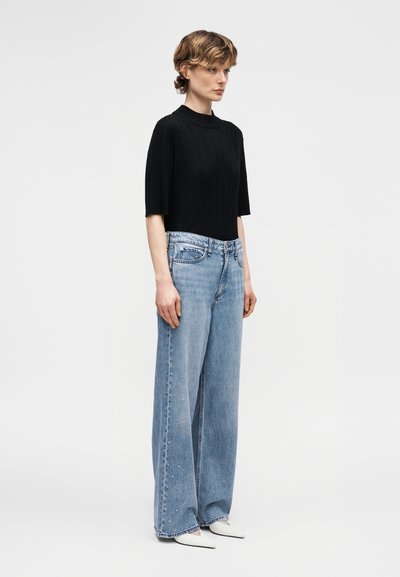 Νεαρό άτομο με κοντά μαλλιά, ντυμένο με μαύρο κοντομάνικο τοπ, ψηλόμεσο φαρδύ τζιν και λευκά παπούτσια με μυτερή μύτη, στεκεται σε πλάγια όψη.