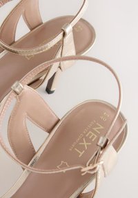 Goldfarbene, metallische Sandalen mit Absatz und pinkfarbener Innensohle, ausgestattet mit kreuzenden Riemen und einem verstellbaren Knöchelriemen, auf einem schwarzen Absatz mit Spitze.