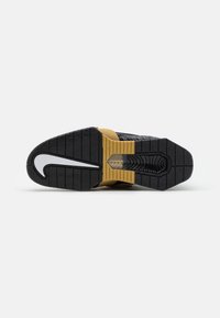 Suola di sneaker nera e oro con motivo testurizzato, caratterizzata da una finestra trasparente e da un distintivo logo a swoosh bianco.