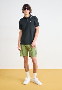 Sort polo-shirt med kontrasterende krave, grøn cargo shorts, hvide sneakers og mørke solbriller. Minimalistisk design med logo detalje.