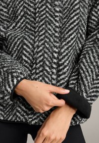 Ręce poprawiające mankiet czarno-szarego, fakturowanego wełnianego płaszcza w jodełkę z miękką, czarną podszewką.