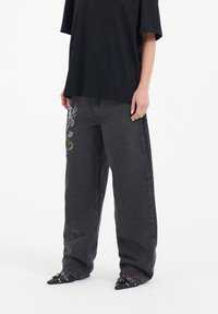 Svart oversized t-shirt parat med högmidjade, löst sittande mörkgrå jeans med färgglad broderad detalj på vänster ben.