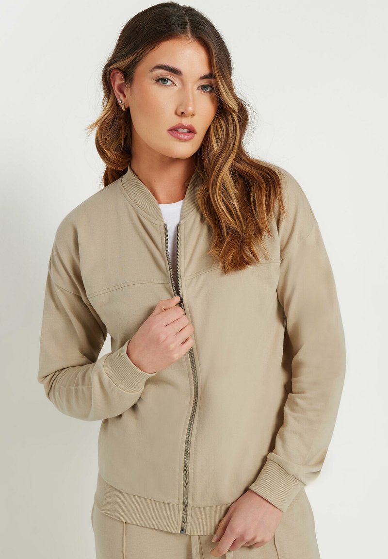 Veste bomber beige zippée avec des poignets et un ourlet côtelés, en tissu texturé et col mandarin, montrée partiellement ouverte.