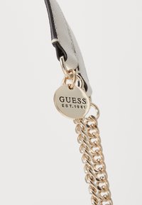 Grå håndtaskerem med en sort accent, der har en guldkæde detalje og et rundt mærke med "GUESS EST. 1981" prægede.