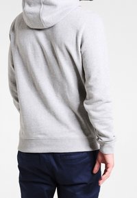 Felpa grigia con cappuccio regolabile, polsini e fondo a costine. Tessuto morbido. Indossata con pantaloni blu navy, mostrando la vista posteriore.