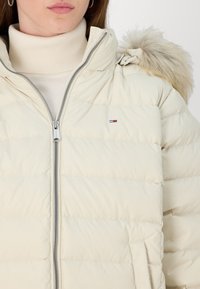Beige dunjakke med hætte, der har pelsdetalje, lynlåslukning og vandret quiltet tekstur. Logo på venstre bryst.
