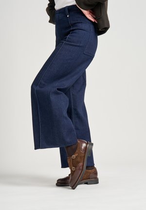 Jeans a gamba larga blu scuro con vita alta, caratterizzati da una tasca posteriore singola e cuciture a contrasto, abbinati a stivali alla caviglia marroni.
