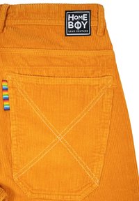 Oranžové kordové nohavice majú na páse nášivku "HOME BOY LOUD COUTURE", s dúhovými pruhmi na vrecku a kontrastným bielym prešívaním.