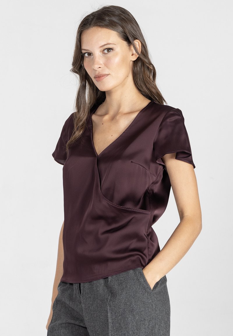 Bordowa satynowa bluzka z krótkimi rękawami, dekoltem w kształcie litery V oraz kieszonką. Miękka tekstura i gładkie wykończenie. Noszona z szarymi, dopasowanymi spodniami.