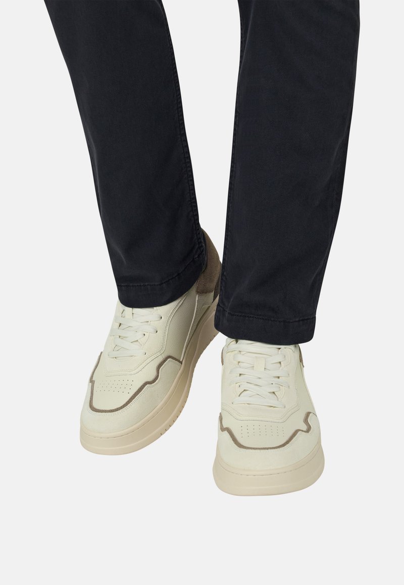 Baskets blanches et beiges à lacets portées avec un pantalon noir, vues de dessus sur un fond clair uni.