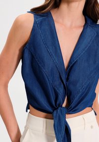 Haut sans manches en denim avec un profond décolleté en V et un nœud à l'avant. Présente une texture lisse et une coupe ajustée en bleu foncé.