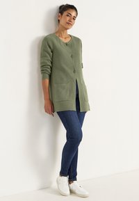 Grön stickad cardigan med rund hals, två framfickor och mörka knappar, kombinerad med mörka skinny jeans och vita sneakers.