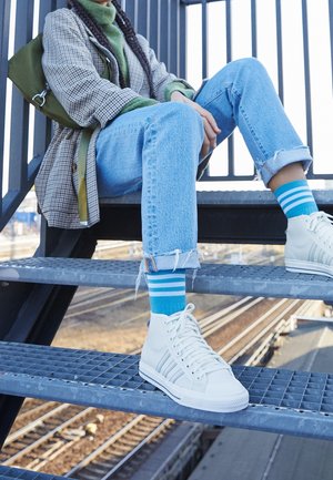 Sneaker bianche alte con strisce blu, jeans di denim e calze a righe turchesi. L'outfit include un cappotto grigio a fantasia e un maglione verde.