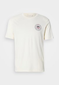 Cremefarvet bomulds t-shirt med rund halsudskæring, korte ærmer og et rundt logo med røde striber og tekst på brystet.