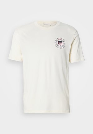 T-shirt en coton couleur crème avec un col rond, des manches courtes et un logo circulaire comportant des rayures rouges et du texte sur la poitrine.