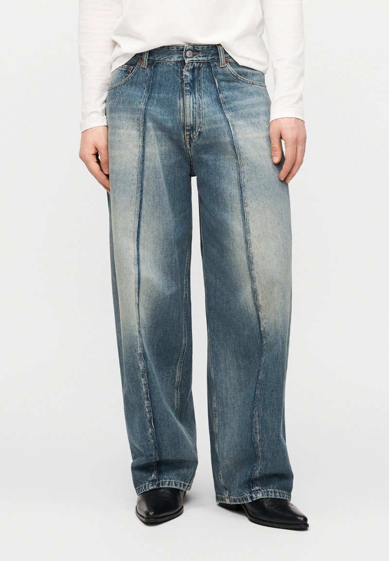 Φαρδιά ξεθωριασμένα μπλε τζιν με λεπτομέρειες ρ αφής στο μπροστινό μέρος, φορεμένα με μαύρα μυτερά παπούτσια και λευκό μακρυμάνικο μπλουζάκι που είναι μπουσουλιασμένο μέσα.