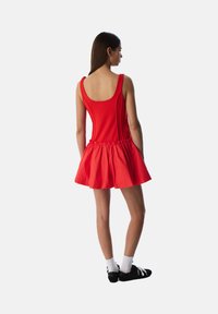 Rode jurk met een getailleerde bovenkant en een uitlopende rok, gemaakt van gladde stof. Heeft een ronde rug en zijzakken, gedragen met zwarte schoenen en witte sokken.