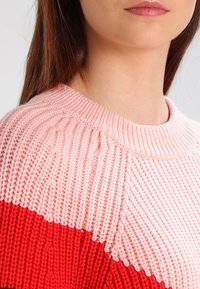 Jeune femme portant un pull en maille bicolore avec des sections rose et rouge, montrant le décolleté et une partie du visage et des cheveux.
