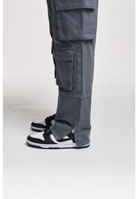 Šedé cargo kalhoty s mnoha bočními kapsami a akcenty na zip, doplněné o černé a bílé tenisky s perforovanými detaily.