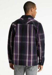 Mann, der rückwärts steht, trägt ein lila und schwarzes kariertes Langarmhemd und hellgraue Jeans auf einem weißen Hintergrund.