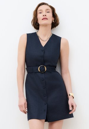 Nainen lyhyillä ruskeilla hiuksilla, joka pukeutunut hihattomaan tummansiniseen napitettuun mekkoon, jossa leveä vyö ja kultaiset korut valkoista taustaa vasten.