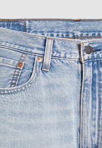 Jean en denim bleu clair en gros plan montrant la taille, la boucle de ceinture, le bouton, la poche avant et les rivets en cuivre avec des détails de couture visibles.