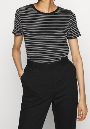 Henkilö, joka pukeutunut mustavalkoraidalliseen lyhythihaiseen paitaan, joka on pujotettu korkeavyötäröisten mustien housujen sisään ja jolla on kädet taskuissa.