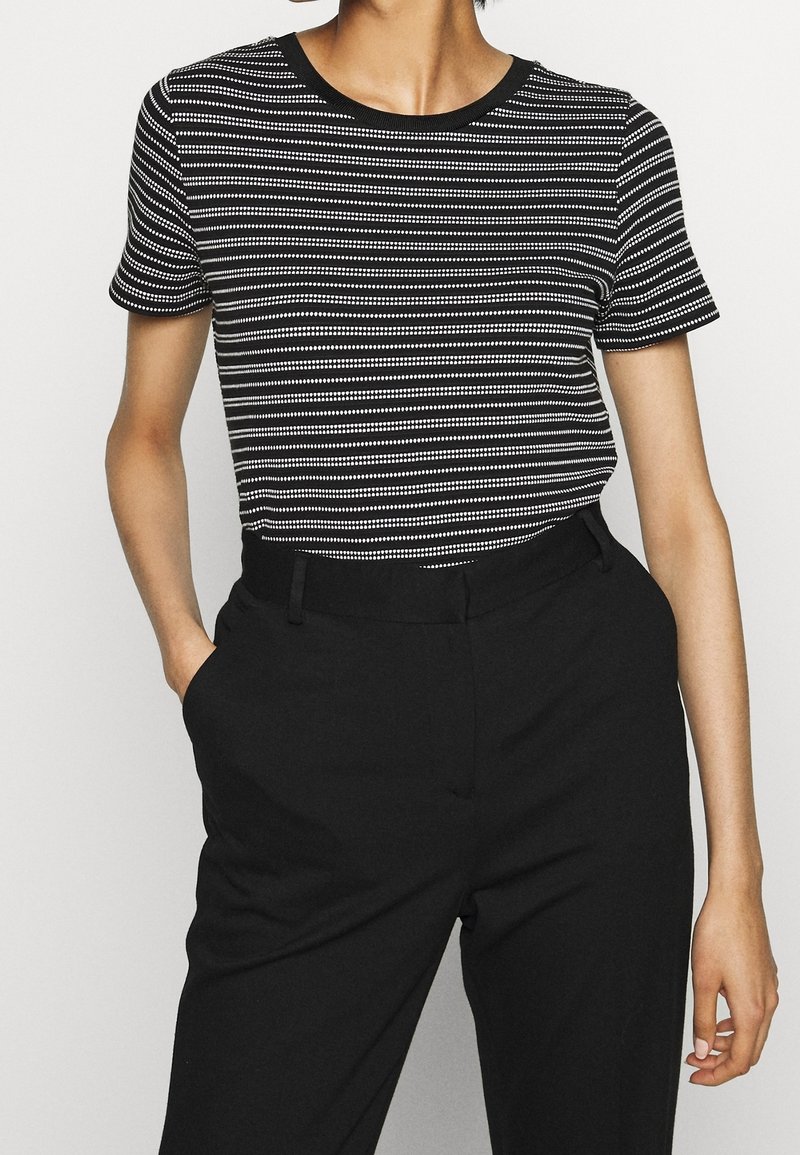 Personne portant une chemise à manches courtes rayée noir et blanc rentrée dans un pantalon noir taille haute, les mains dans les poches.