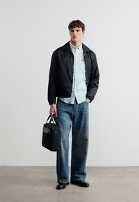 Νέος άνδρας στέκεται κρατώντας μια μαύρη τσάντα, φοράει μαύρο σακάκι, ανοιχτό μπλε πουκάμισο με κουμπιά, άνετο μπλε τζιν και μαύρα παπούτσια.