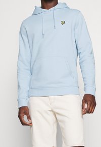 Sudadera azul claro hecha de una mezcla de algodón, con un bolsillo tipo canguro, puños acanalados y un pequeño logo negro y amarillo en el pecho.