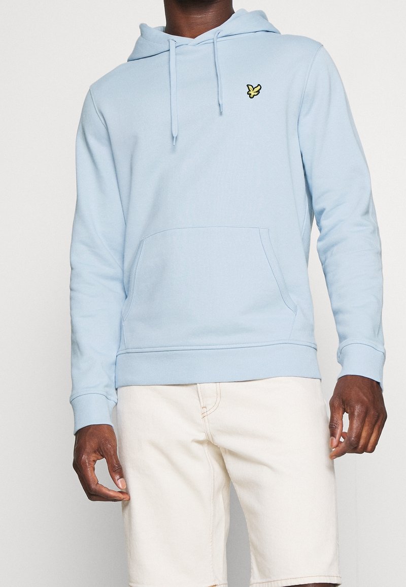 Sudadera azul claro hecha de una mezcla de algodón, con un bolsillo tipo canguro, puños acanalados y un pequeño logo negro y amarillo en el pecho.