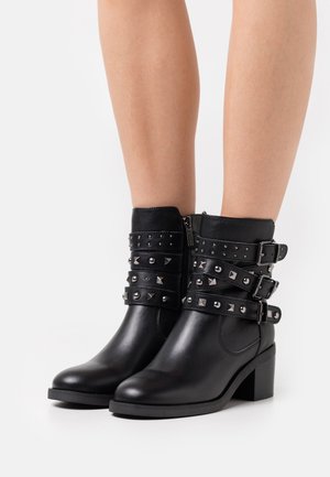 Bottines en cuir noir avec un bout carré, un talon moyen et des sangles cloutées, dotées de clous en forme de pyramide et ronds en argent, avec un zip latéral.