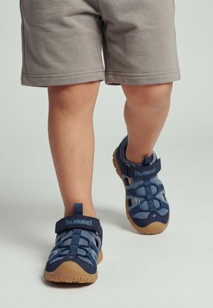 Barn iført grå shorts og blå lukkede sandal med velcrolukning går på en lys grå gulv.