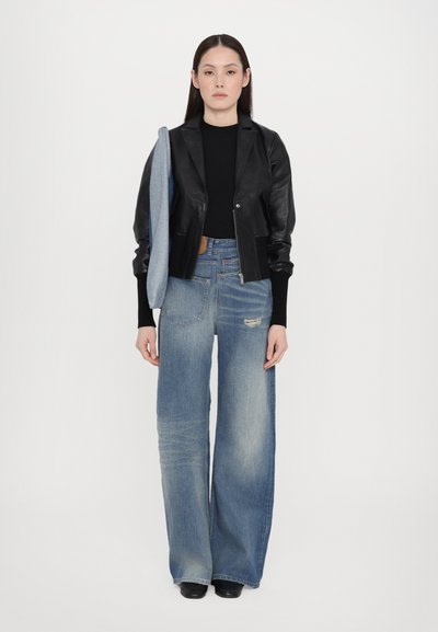 Black leather cropped jacket over a black top, paired with wide-leg light blue jeans with distressed details. Held denim accessory. Black shoes.