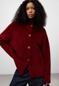 Maglione oversized bordeaux con una finitura morbida e testurizzata, caratterizzato da un alto colletto e sei bottoni tondi lungo la parte anteriore.