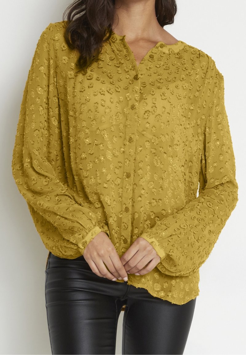 Femme aux cheveux foncés portant un chemisier à manches longues texturé jaune moutarde avec des boutons et un pantalon en cuir noir, mains jointes à la taille.