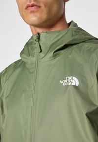 Veste à capuche vert olive en tissu lisse et léger, équipée d'une fermeture éclair à l'avant et d'un logo blanc sur la poitrine.