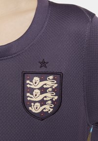 Jersey viola con texture, caratterizzato da un'emblema a scudo con tre motivi a leone e accenti di rosa, sormontato da una stella sopra il crest.