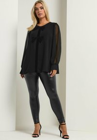 Schwarze Bluse mit durchsichtigen langen Ärmeln, einem zentralen Samtband und zwei Schleifen. Kombiniert mit glänzenden schwarzen Leggings und schwarzen Riemchen-Heels.