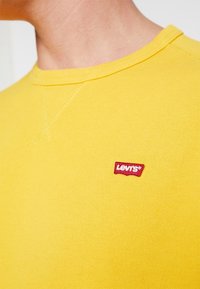 Sweatshirt jaune en maille avec col rond et coutures visibles. Présente un petit patch logo Levi's rouge sur la poitrine.