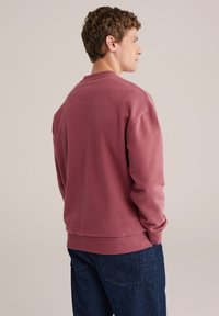 Temno rdeč pulover iz mehkega materiala, z okroglim izrezom in dolgimi rokavi. Ima rebraste manšete in rob, kombiniran z temno modrimi kavbojkami.