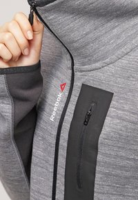 Giacca sportiva grigia con colletto alto, tasca con zip nera e texture sottile. Presenta il logo Reebok in rosso sul davanti.