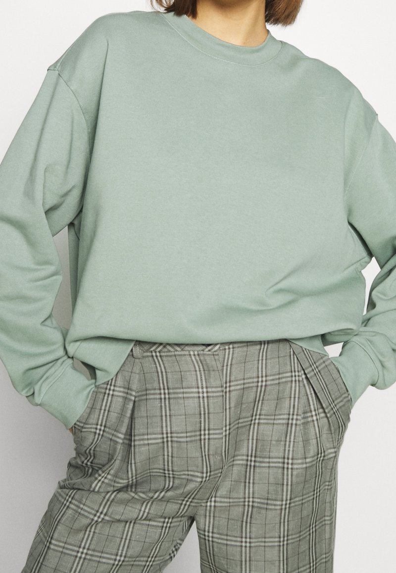Person wearing light green oversized sweatshirt and high-waisted gray plaid trousers, hands in pockets, against plain background.