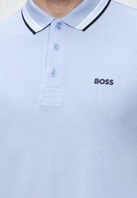 Polo bleu clair avec col rayé blanc et noir, trois boutons et logo "BOSS" brodé sur la poitrine.