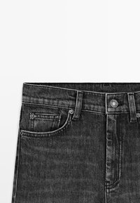 Primo piano della vita di jeans in denim neri che mostra le tasche frontali, gli anelli per la cintura, la chiusura con bottone e le cuciture dettagliate su sfondo bianco.