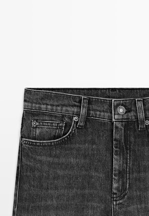 Primo piano della vita di jeans in denim neri che mostra le tasche frontali, gli anelli per la cintura, la chiusura con bottone e le cuciture dettagliate su sfondo bianco.